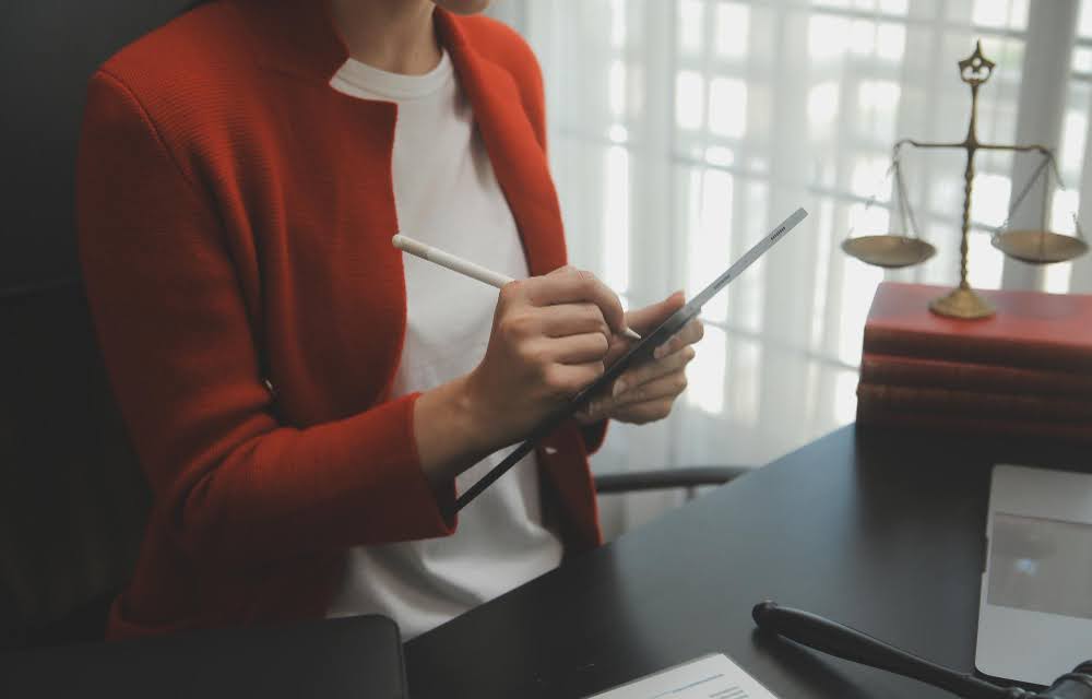 Woman in red jacket taking notes on her tablet while sitting in front of desk