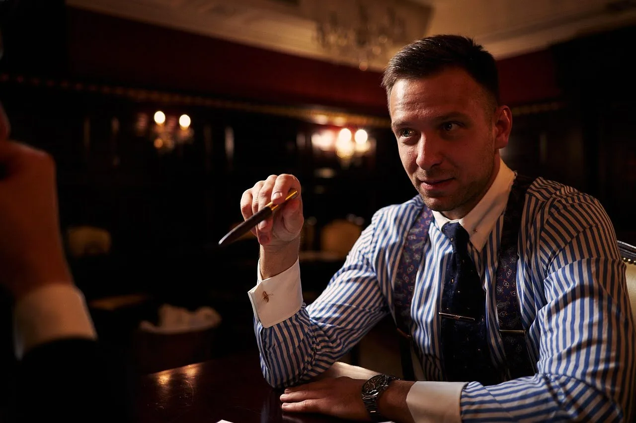 A well-dressed man in a striped shirt and tie sits at a polished wooden table in a dimly lit room, holding a pen and appearing engaged in conversation, possibly during a legal deposition.