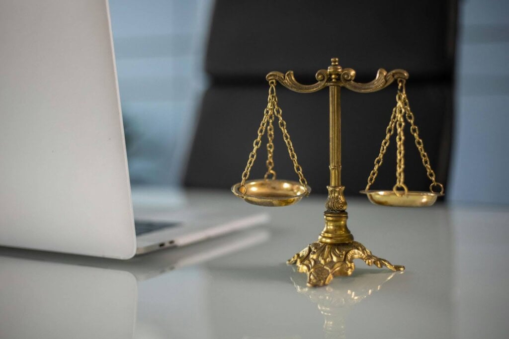 A golden balance scale placed on a desk beside an open silver laptop, symbolizing justice and law in a modern office setting