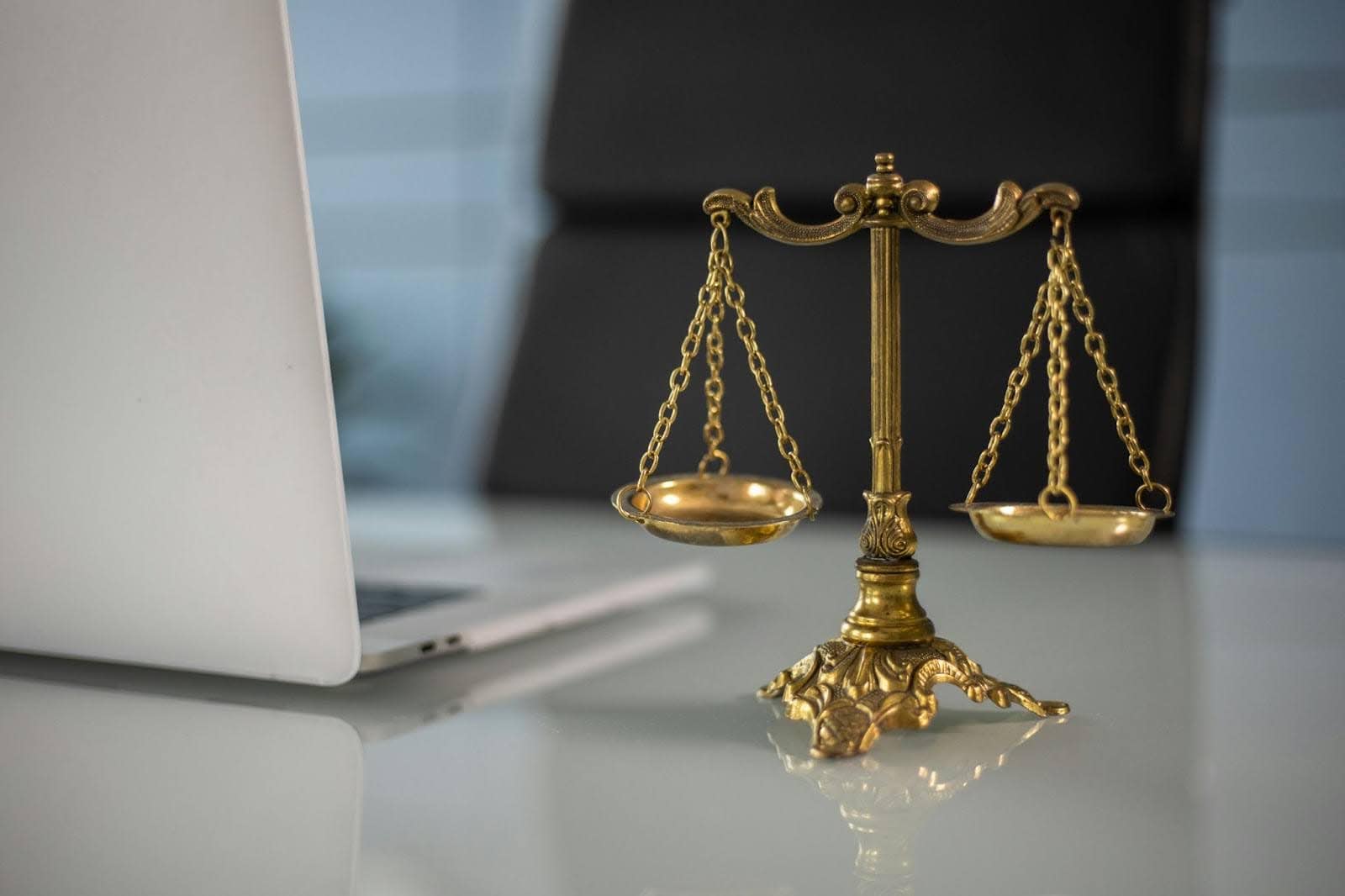 A golden balance scale placed on a desk beside an open silver laptop, symbolizing justice and law in a modern office setting